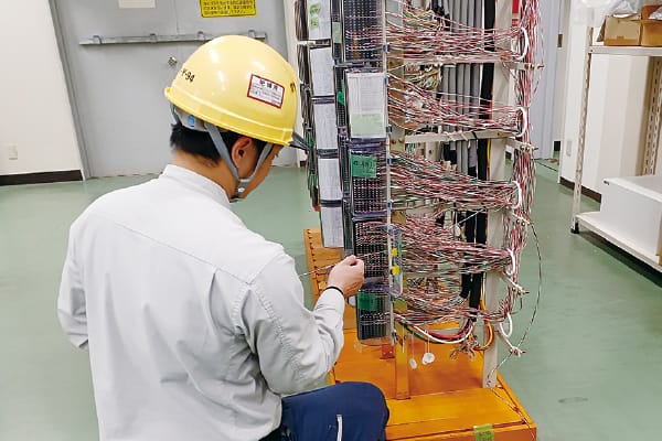 屋内で作業員が多数の配線が施された制御盤を点検している様子